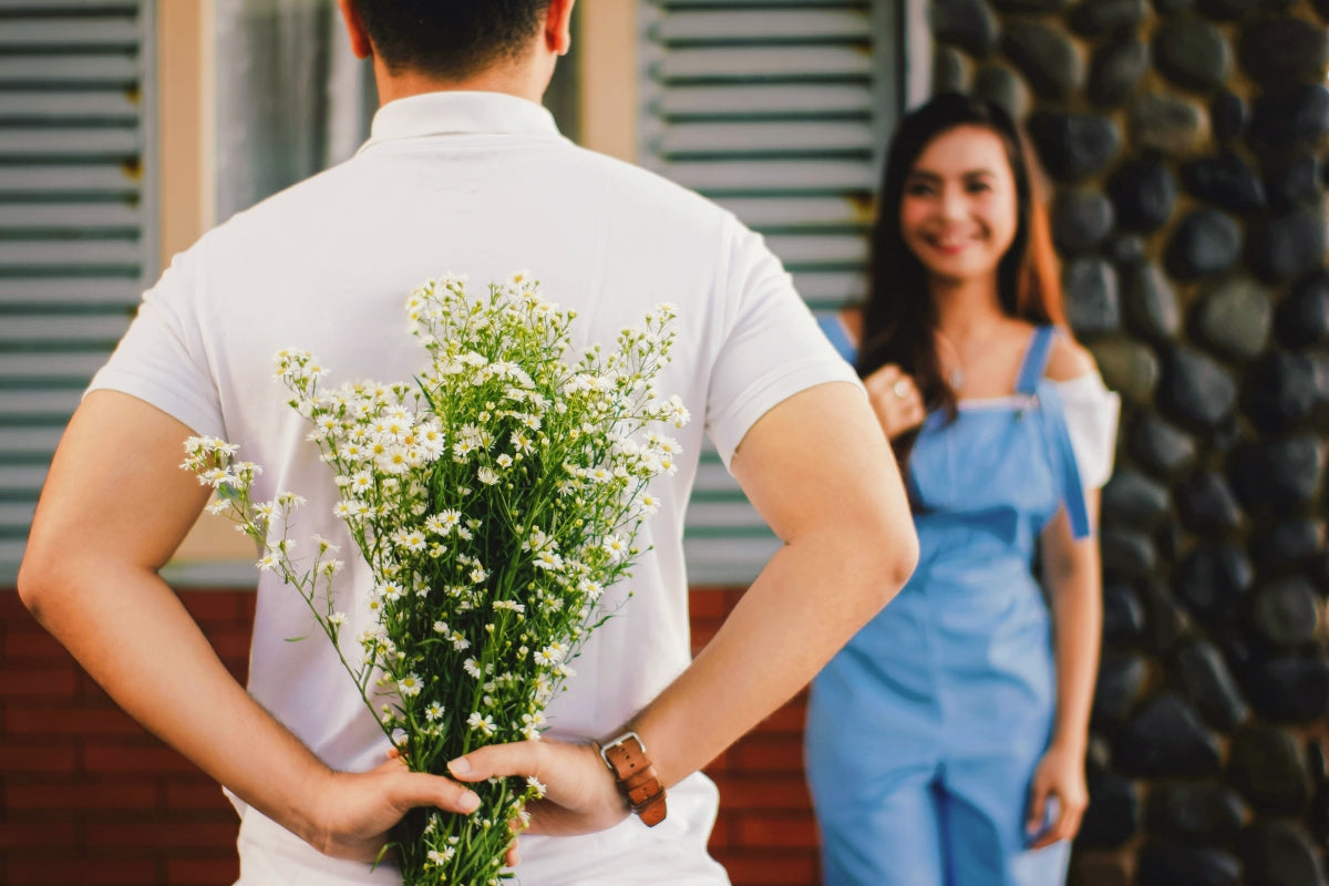 A man is holding flowers behind his back and preparing to surprise his girlfriend